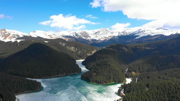 Aerial view of a frozen mountain lake surrounded by forest and snowy mountains in spring season. alt
