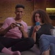Young Woman Surfing Internet Using Mobile Phone and Her African American Boyfriend Playing Video