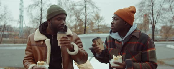Two Black Men Having Snack with Coffee and Chatting by Car alt