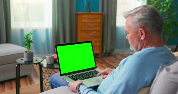Older Man at Home Sitting on a Couch Works on a Laptop Computer with Green alt