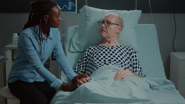 Sick Elder Patient Laying in Bed Talks to African American Visitor alt