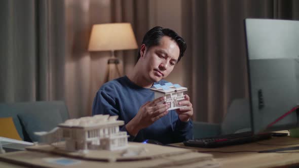 Asian Male Engineer Holding And Looking At House Model With Solar Panel While Working On Desktop alt