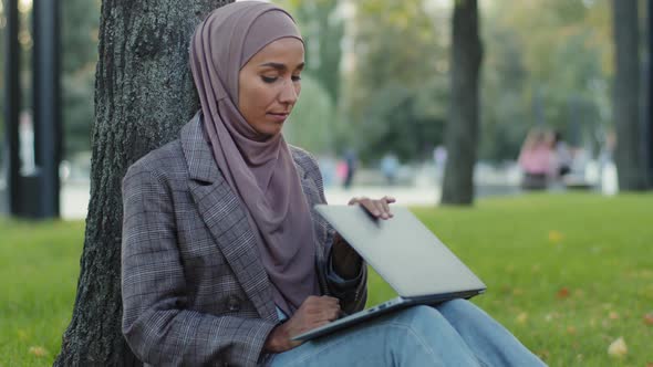 Tired Girl Student in Hijab Young Business Woman with Laptop Sitting in Park Near Tree on Green Lawn alt