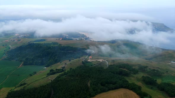 Flying over the clouds that shroud a single house on top of a hill surrounded by lush green meadows alt