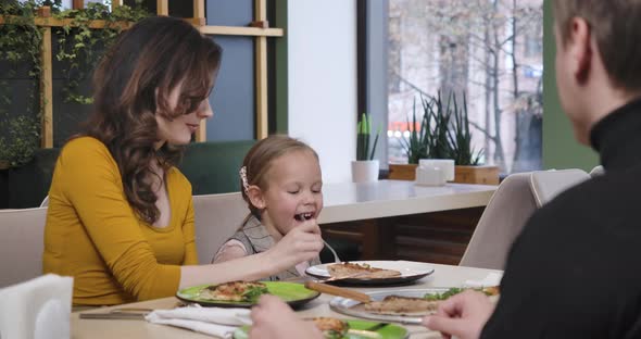 Happy Young Mother Feeding Cute Charming Daughter with Pizza in Restaurant alt