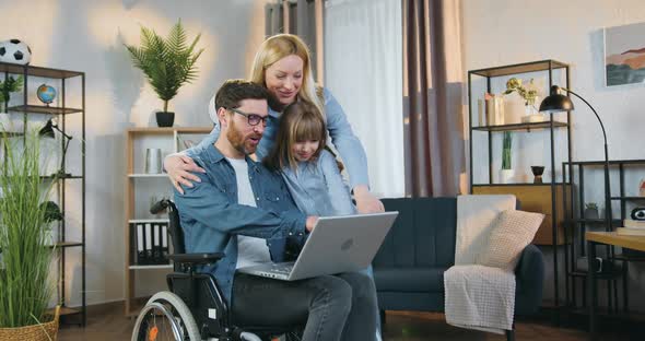 Man in Wheelchair Applying Laptop Together with His Daughter and Pretty Joyful Wife at Home alt