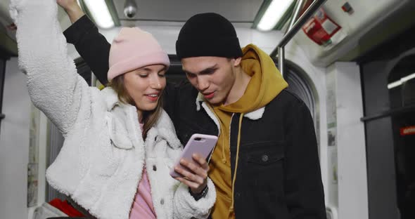 Cheerful Millennial Couple Looking at Smartphone While Going on Subway. Young Pretty Girl Holding alt