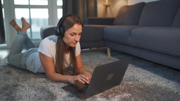 Woman Is Lying on the Floor, Working on a Laptop and Listens To Music on Headphones alt