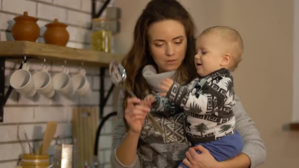 Pretty Young Woman with a Baby in Her Arms in Modern Kitchen Preparing Breakfast alt