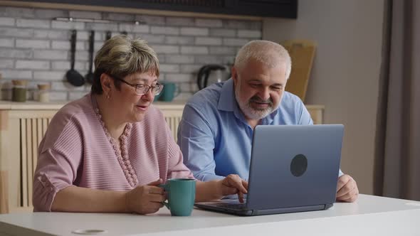 Old Man and Woman are Learning to Use Laptop Together Sitting at Home Kitchen at Weekend Pressing alt