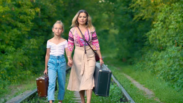 Serious Mom and Daughter Holding Hands Walking on Railway with Traveling Suitcase alt