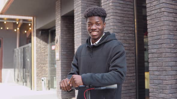 A Young African American Man in Casual Dark Clothes with a Scooter Stands Near a Building with a alt