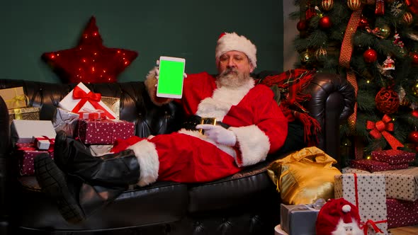 Santa Claus Lies on the Sofa Near Decorated Christmas Tree and Gift Boxes Shows Tablet with a Green alt