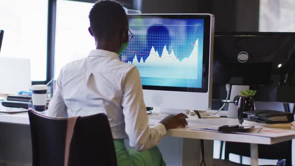 African american woman wearing face mask using computer while sitting on her desk at modern office alt