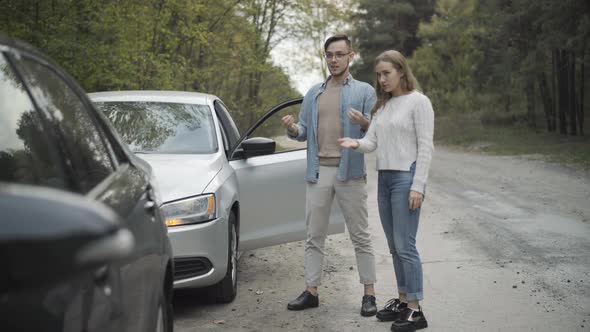 Wide Shot of Young Caucasian Man and Woman Talking and Gesturing Looking at Broken Cars on Roadside alt
