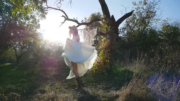 A young beautiful brunette in a white dress rotates and waves her dress on a meadow in the forest. S alt