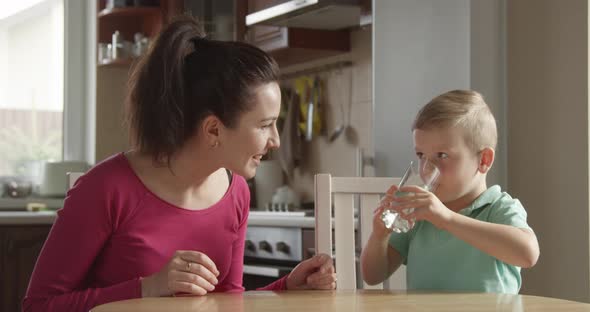 Kid Is Drinking A Glass Of Water With Mother Sitting Close By At The Kitchen alt