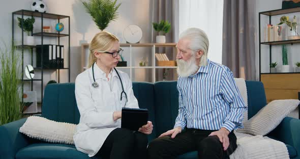 Woman-Doctor Sitting with Old Bearded Man on the Couch in His Home and Telling alt
