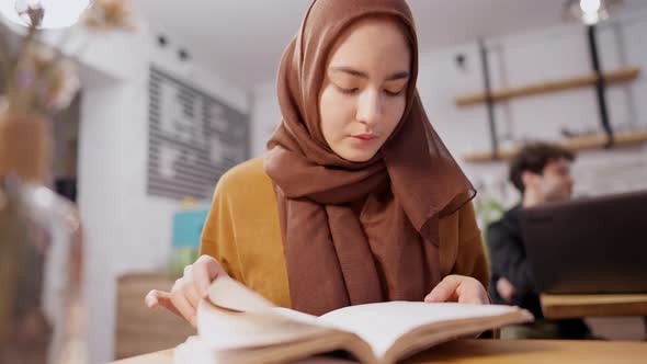 Absorbed Slim Middle Eastern Young Woman Turning Pages of Book Sitting in Cafe alt