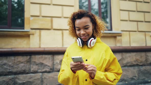 Happy African American Student Is Using Smartphone Browsing or Texting Friends Standing Outdoors in alt