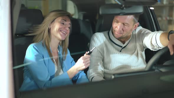 Panning Shot of Cheerful Couple Talking Sitting in New Automobile with Car Key Smiling alt