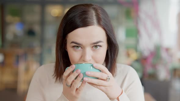 Brunette Caucasian Young Woman with Hazel Eyes Drinking Coffee and Looking at Camera alt
