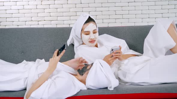 Two happy Asian girls in white bathrobes with facial mask using smart phone on sofa. alt