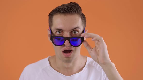 Excited Hipster Man in Surprise Shoots Sunglasses Celebrating Success in Studio on Orange Background
