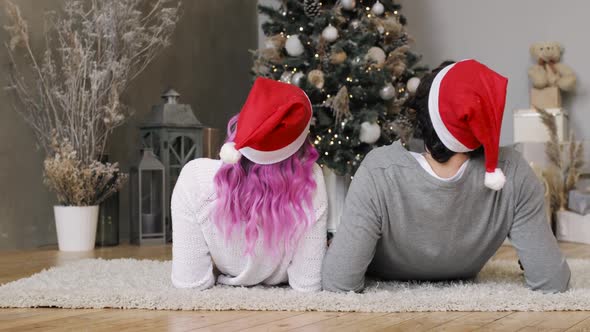 Man and Woman in Santa's Red Hats Lying Together Near Christmas Tree, Back View.