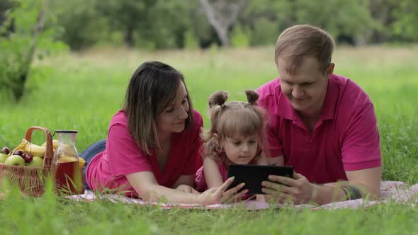 Family Weekend Picnic. Daughter Child Girl with Mother and Father Play Online Games on Tablet alt
