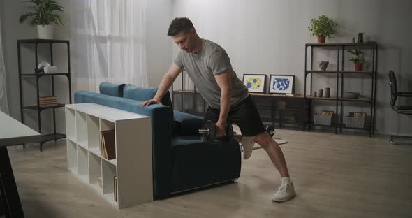 Powerlifter Is Doing Exercise with Dumbbell at Home Leaning on Couch in Living Room Adult Sporty Man alt