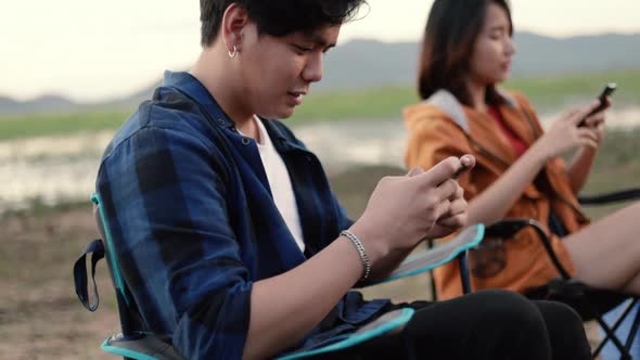 Asian man using smartphone playing games with a girlfriend sitting beside a tent camping.