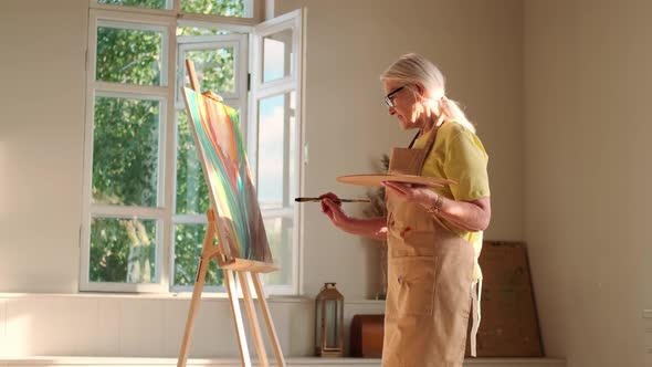Senior Woman Artist Paints a Picture on Canvas alt