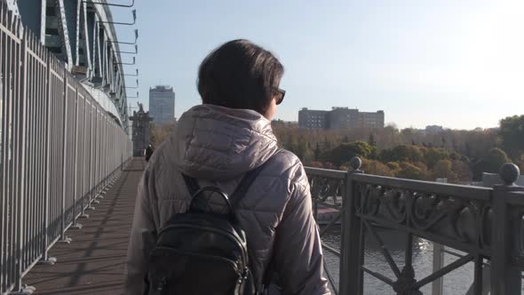 A Middleaged Woman Walks Alone with a Bouquet of Yellow Leaves on a Bridge Over a River on a Sunny alt