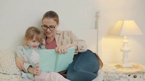 Caring Mother Reading Book To Adorable Little Girl Sitting in Bed in Bedroom Together alt