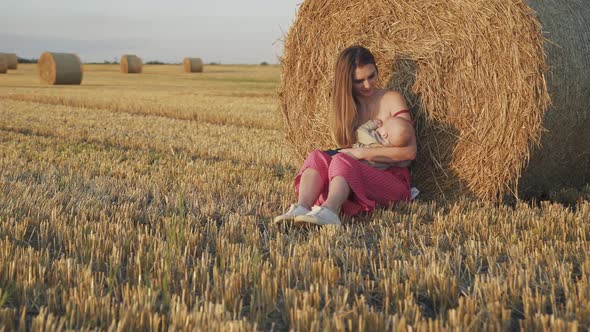 Beautiful Young Mother Feeds Baby Son with Breast at Haystack in Summer Field alt