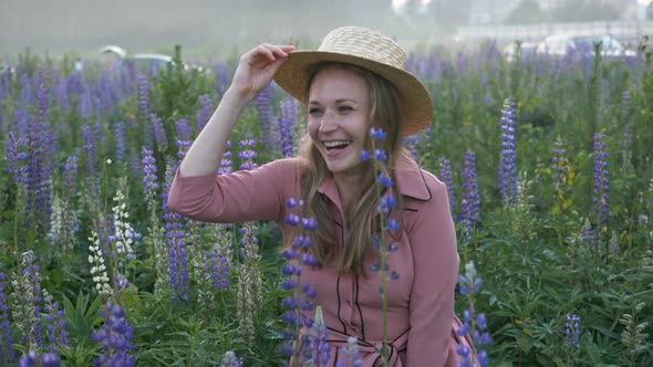 Girl in Pink Dress Rises From Grass in Blooming Lupine Field and Waving Boater with Straw Hat at alt