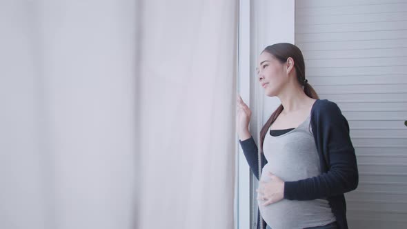 Happy Asian pregnant woman standing looking out the window at home. alt