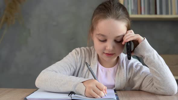 Schoolgirl Girl Talking on the Phone and Making Notes in a Notebook Sitting at Home at the Table alt