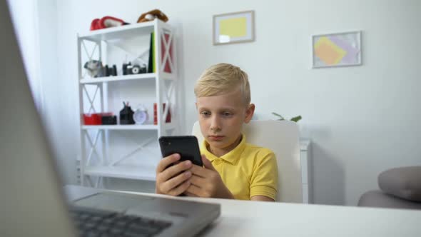 Little Male Kid Sitting in Front of Laptop and Watching Video on Smartphone, App