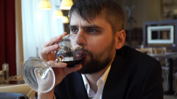 Young Serious Businessman with Beard Sitting in Restaurant and Drinking Red Wine From Glass