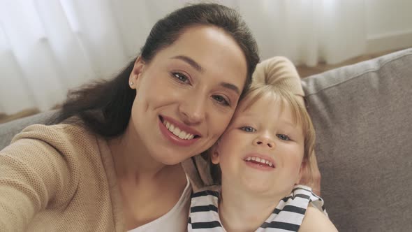 Young Mother and Small Preschooler Daughter Look at Camera Making Selfie and Smiling at Home alt