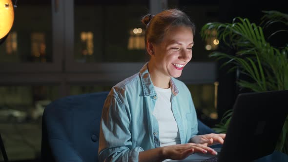 Woman is Sitting in the Armchair and Working on a Laptop at Night or Texting Someone alt
