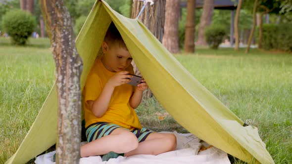Young Traveler in Tent Playing Video Game. Traveling Concept. Boy in Hand Made Tent Plays Game on