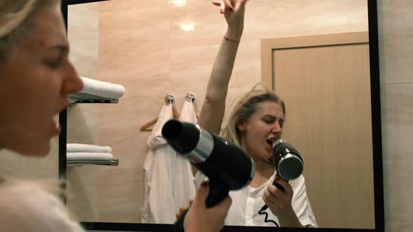 Young Woman Sings in a Working Hair Dryer Opposite the Mirror Standing in the Bathroom