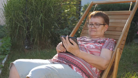 An Elderly Woman with Glasses Reads an Ebook Sitting on a Wooden Chaise Longue in the Backyard of a