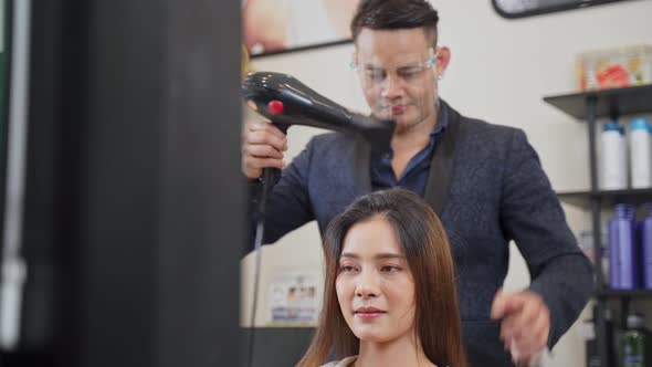 Professional male stylist wearing face shield using hair dryer blow woman's hair in beauty salon. alt