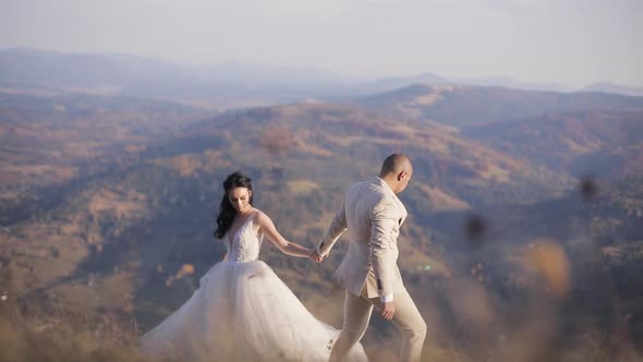 Beautiful and Lovely Bride in Wedding Dress on the Mountains in Sunny Autumn Day. Groom Infront Her.