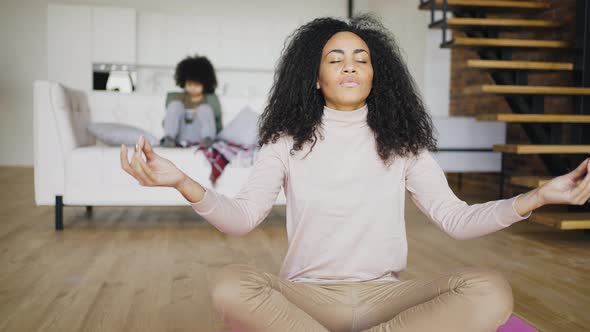 Calm Young Mixed Race Mother Meditates at Home While Her Son Plays with Smartphone alt