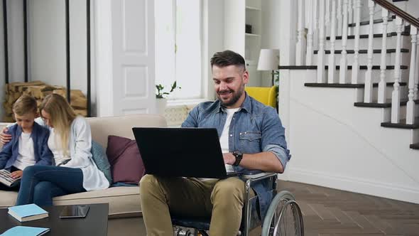 Bearded Guy in Wheelchair Working on Laptop and His Wife and Son Reading Book on the Sofa Near Him alt
